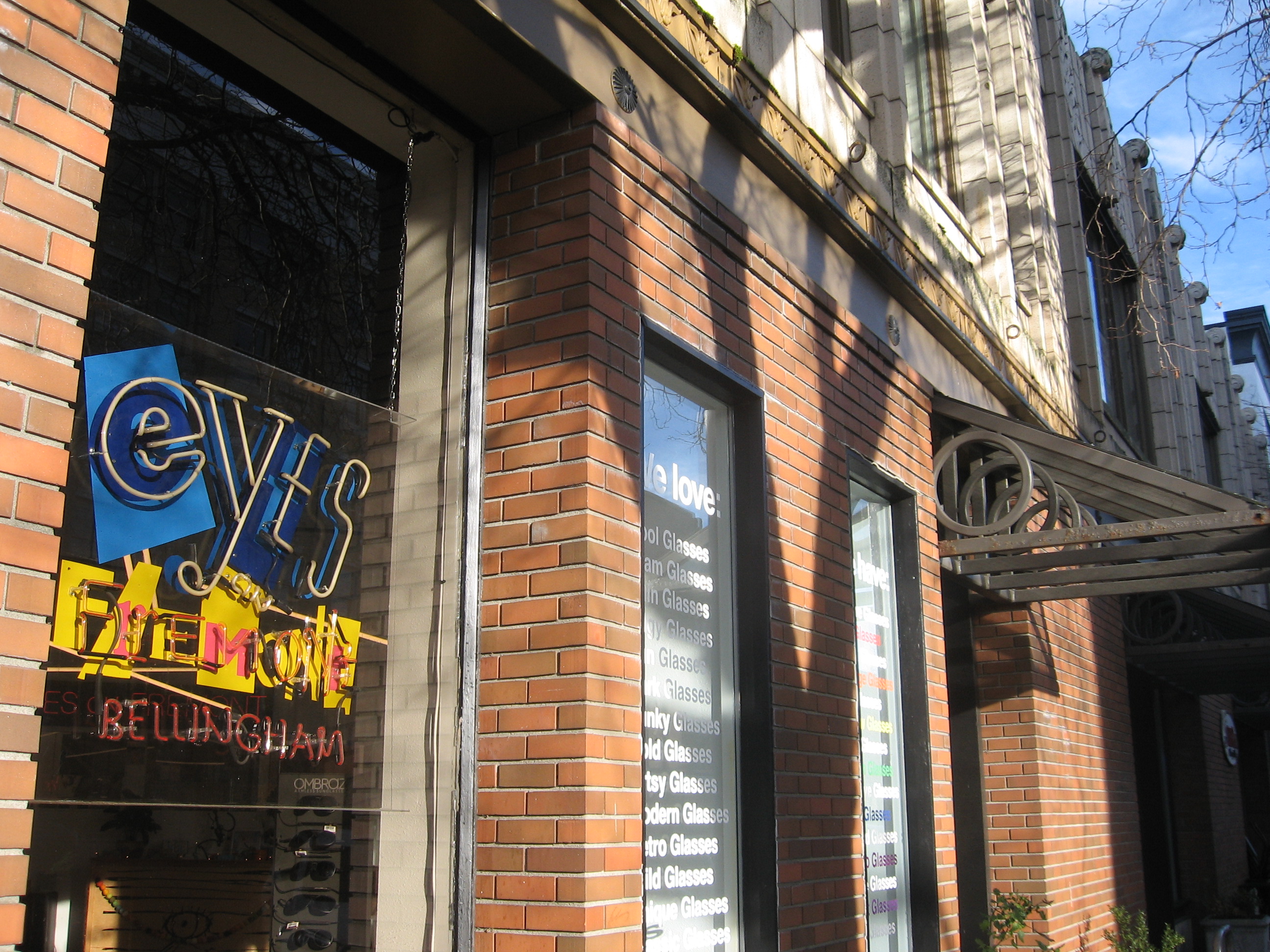 Daylight photo of a funky sign for 'Eyes on Fremont, Bellingham' eyewear store. In the window next to it there's a list of all sorts of eyeglass styles.